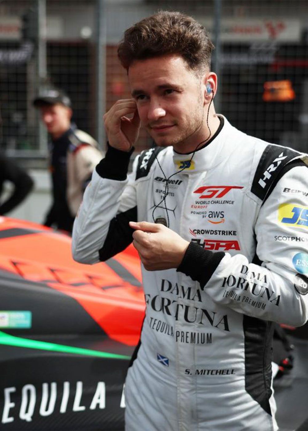 Driver in a white race suit fitting an earpiece in the pit lane